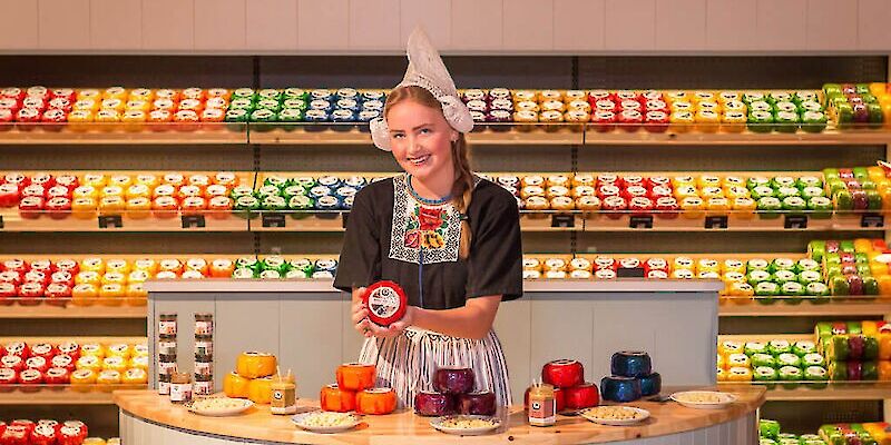 Woman in traditional Vrouw Antje clothing behind a counter with various cheeses, with a wall of color-sorted mini cheeses behind her.