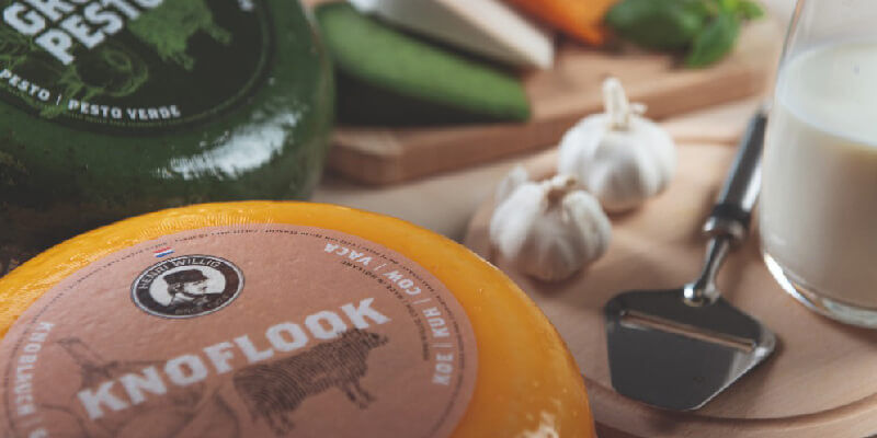 Two cheese wheels partially in view with a glass of milk, a cheese slicer and two bulbs of garlic on a wooden board.